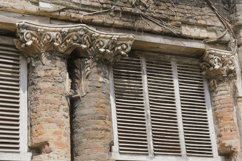 detailed construction features intricate corinthian capitals on twin brick pillars, sunburst relief motifs, and wooden louvered shutters set in decaying masonry