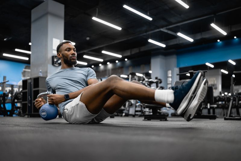Fit man performs kettlebell Russian twist on gym floor, engaging core muscles for strength, balance and endurance during an intense workout session in a modern fitness center.