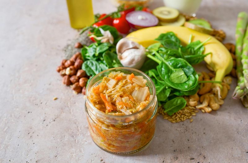 Foreground focus on a jar of kimchi surrounded by leafy greens, banana, grains and nuts, symbolizing gut nourishment and internal harmony. Soft neutral background provides usable copy space, ideal for editorial and commercial storytelling about microbiome health and resilience.