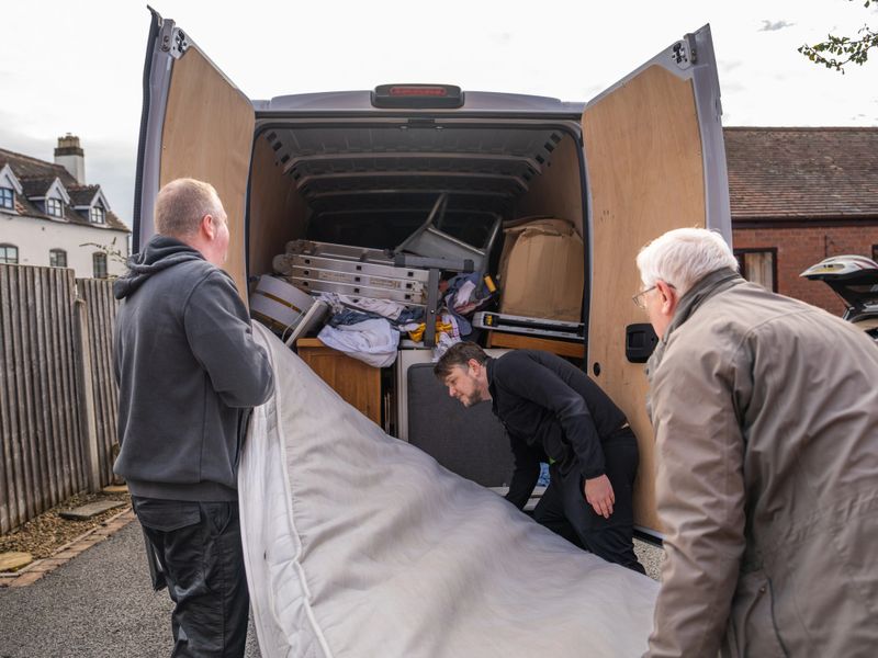 Three men working together to lift and load a mattress into a removal van outside a residential building during a house move. Concept of relocation, teamwork and manual work on moving day.