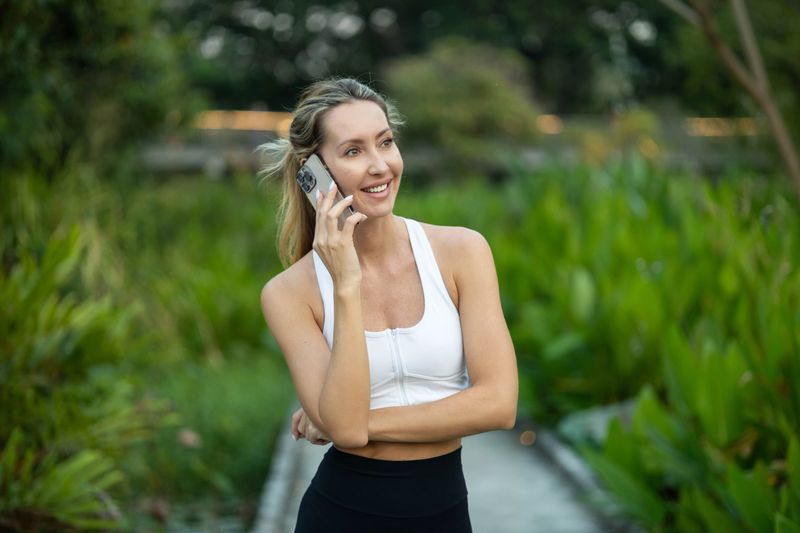 A cheerful woman speaks on her phone, enjoying a green pathway and adding positive vibes to her day.