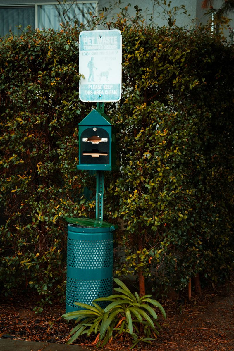 A green pet waste station featuring a dispenser for bags and a perforated trash receptacle stands against a backdrop of lush, dense green bushes and trees