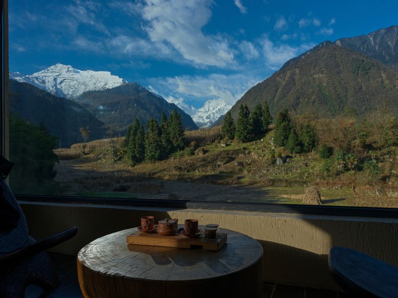 Traditional tea set placed on a wooden table inside a mountain lodge, overlooking snow-capped peaks and forested valley through a panoramic window. Peaceful travel and luxury retreat atmosphere.