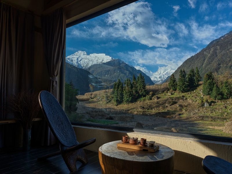 Traditional tea set placed on a wooden table inside a mountain lodge, overlooking snow-capped peaks and forested valley through a panoramic window. Peaceful travel and luxury retreat atmosphere.