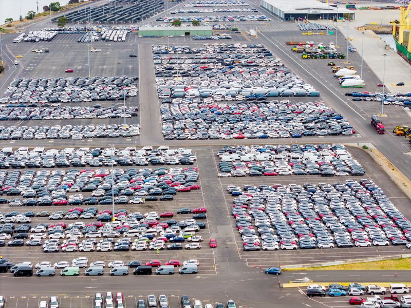 Thousands of new cars, vans, and SUVs are parked in neat rows at a vast port terminal, with a cargo ship docked nearby.