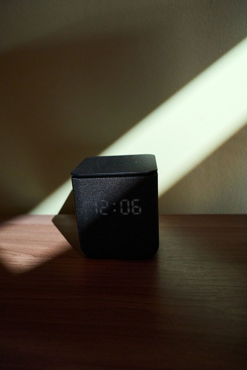 A sleek black smart speaker sits on a wooden surface, bathed in soft sunlight creating a dramatic shadow effect. Represents time, technology, and modern design
