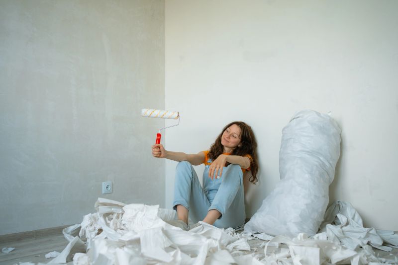 Young woman taking a break during apartment renovation, holding a paint roller and sitting on the floor among old wallpaper, enjoying the progress of her diy project