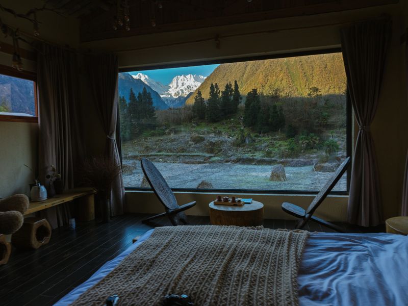 Cozy cabin bedroom interior with bed, lounge chairs, and a panoramic picture window overlooking a forest valley and snowy mountains in early morning light, Kangding
