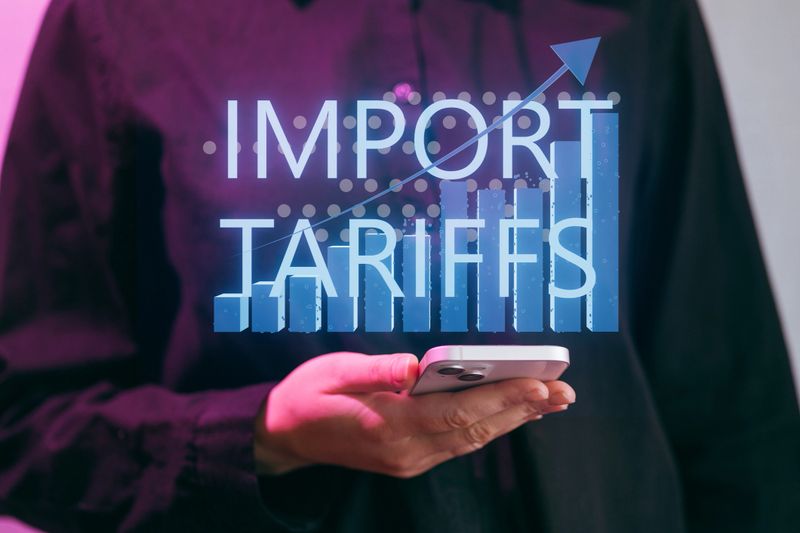 A person holds a smartphone displaying a graphic on import tariffs. The graphic shows an upward trend in trade metrics. The setting appears to be an office or workspace.
