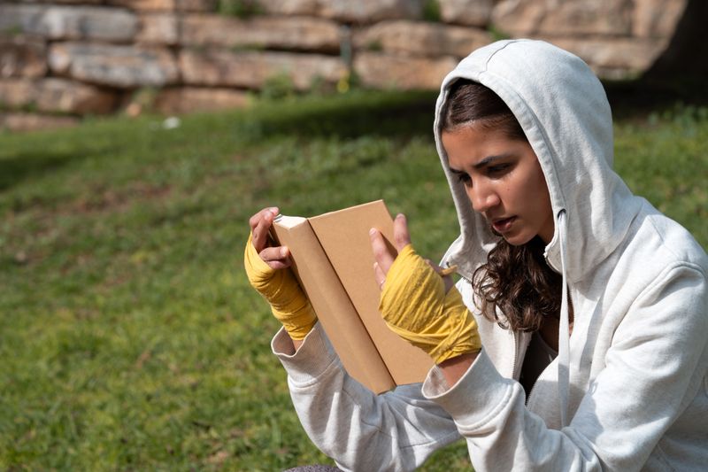 Young woman in a hoodie with yellow hand wraps focused on reading a book outdoors on a sunny park lawn, contemplative and strong