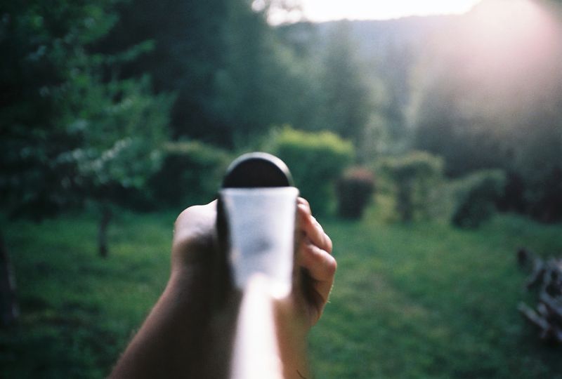 Authentic 35mm film scan with a POV perspective of aiming an air gun in a sunny garden. Features dramatic sun flare, green backyard, and film grain.
