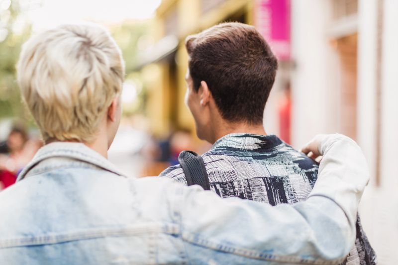 Rear view of two friends with arms around each other walking down a colorful city street, enjoying a casual day out together.