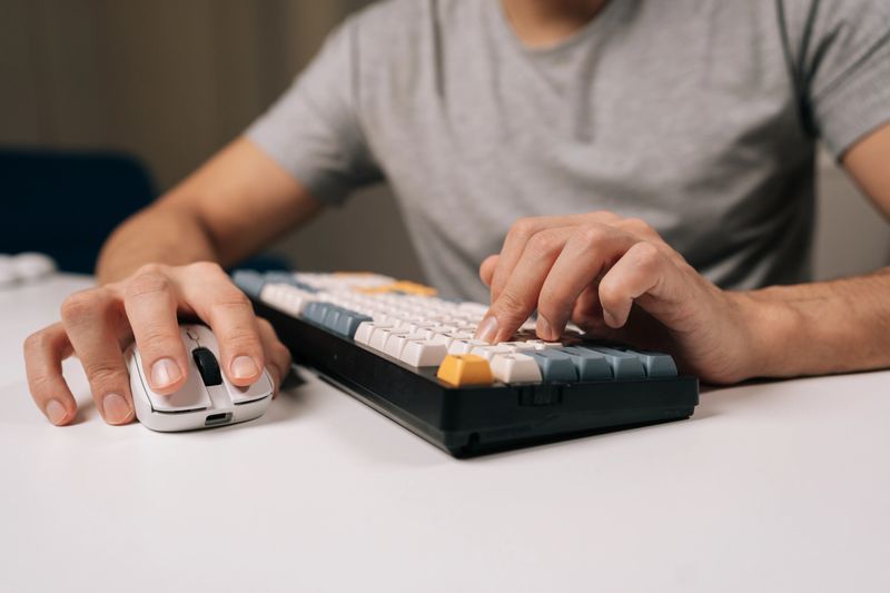 Close-up cropped shot of hands typing on colorful mechanical keyboard next to white wireless mouse on clean desk, conveying gaming, coding, remote work, and modern professional computing setup.