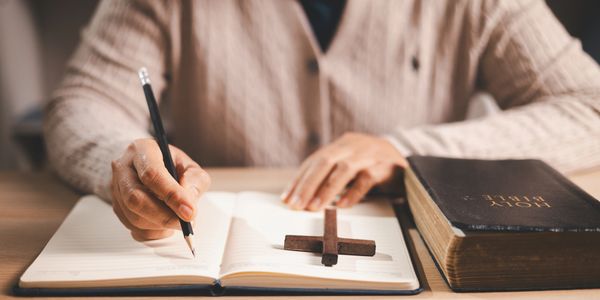 Person writing in a notebook with a cross and Bible nearby.