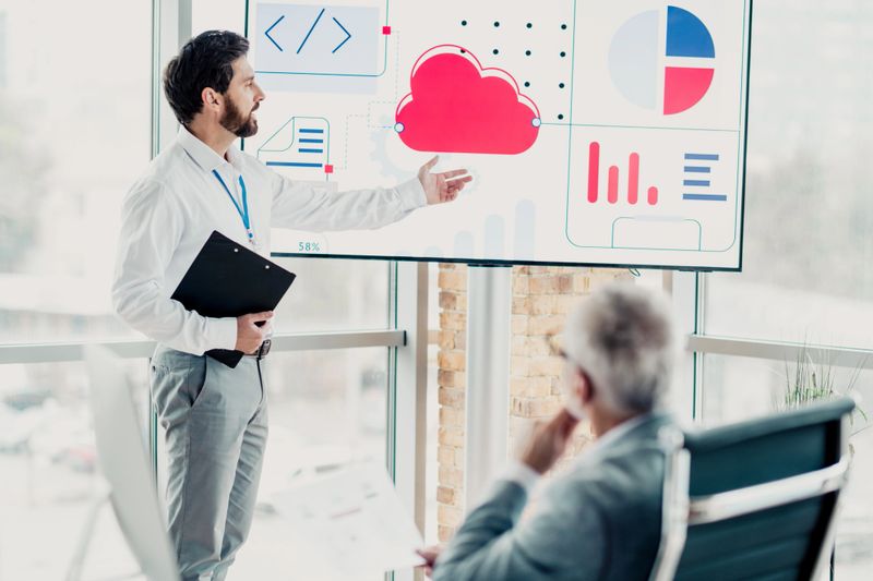 A professional male presenter explains data on a large screen in a bright loft office while an audience member observes. The scene conveys corporate, team, and strategic collaboration and modern work culture.