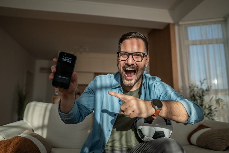 Enthusiastic man with a soccer ball cheering and pointing at a smartphone while celebrating an online sports betting victory or staying updated on a live game from his comfortable home sofa