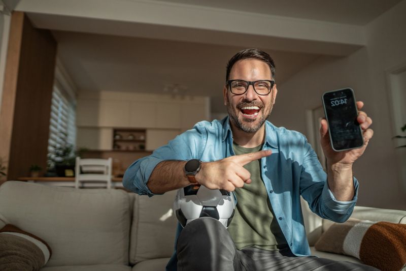 Man celebrating a winning online football bet, sitting on a living room couch holding a soccer ball and smartphone, smiling broadly and pointing at the screen in daylight
