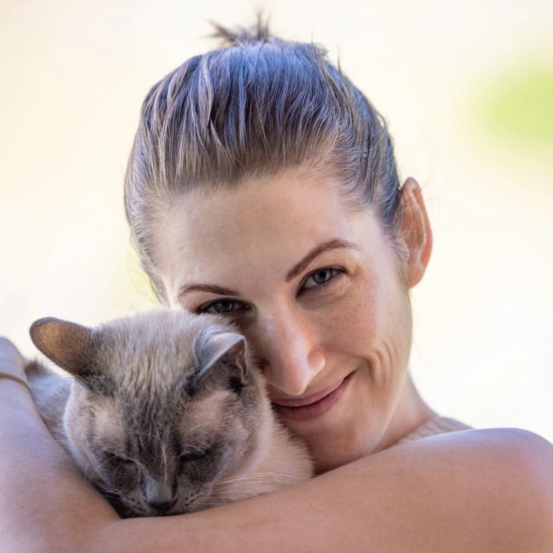 A close-up portrait of a smiling woman embracing her cat, conveying warmth, affection and a strong pet-owner bond in soft natural light. Intimate, cozy and tender moment between human and animal.