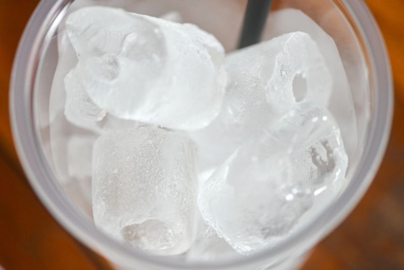 Close-up top view of ice cubes in a clear plastic cup with a straw, bright natural lighting, wooden surface background