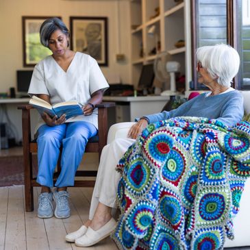A caregiver reads a book to an elderly woman wrapped in a colorful blanket.