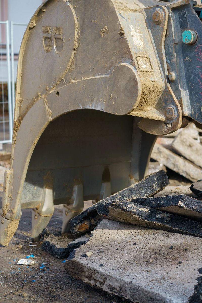 Close-up of hydraulic excavator bucket tearing asphalt and concrete during demolition works. Heavy machinery at urban construction site.