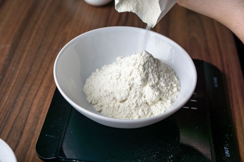 Close-up of flour being poured into a white bowl on a digital kitchen scale. Careful measurement of dry ingredients during home baking preparation on a wooden countertop.