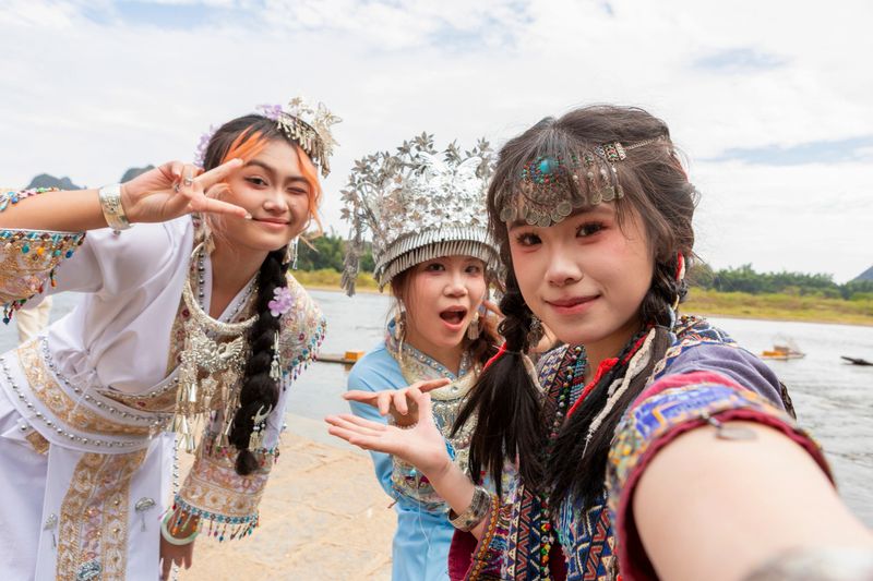 Three young Chinese women in ornate Hmong traditional costumes and silver headdresses smiling and taking a selfie by a scenic Guilin river, celebrating friendship and culture