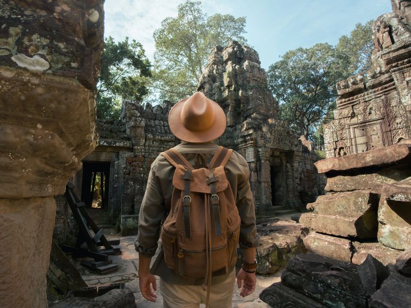 Male adventurer wearing a hat and backpack walks into the ruins of Ta Som Temple, Angkor Wat. Great for Southeast Asia travel, cultural exploration, or heritage tourism promotion.