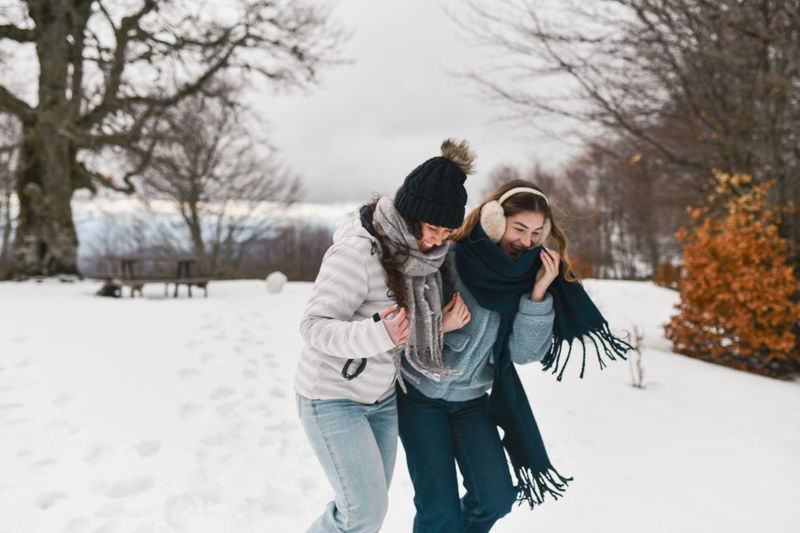 Female friends dressed warmly for winter walk together in snowy surroundings, smiling as they embrace the chilly weather.