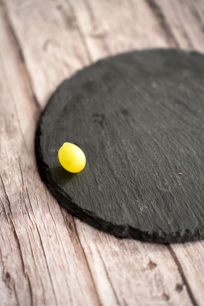 close-up of a single yellow grape on the edge of a round black slate coaster resting on weathered wooden planks, minimalist rustic tabletop scene with soft natural light and textured surfaces