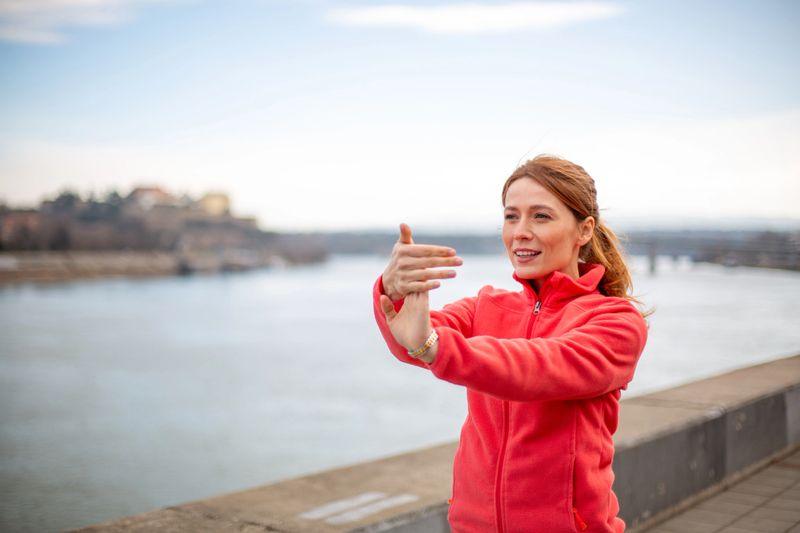 Woman stretching hands and wrists by river, preparing for active outdoor workout