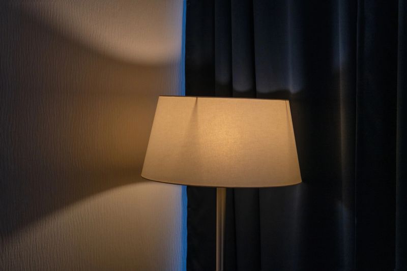 A lit floor lamp with a warm beige lampshade creates a cozy, intimate mood. Soft light illuminates a textured wall and contrasts with dark, heavy curtains in a quiet room, creating deep shadows