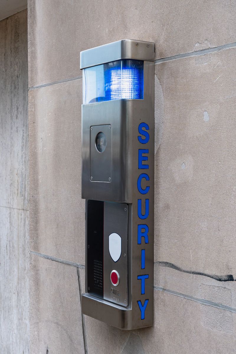 Modern security intercom panel mounted on a stone wall. Stainless steel emergency call box with surveillance camera, speaker, red call button and blue flashing light on top. The word “SECURITY” is displayed vertically on the side. Concept of safety, surveillance, access control and building protection