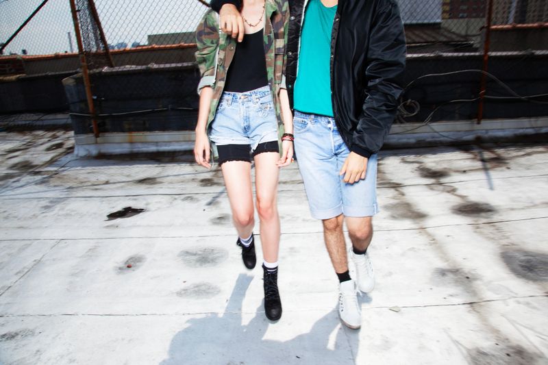 Three young friends stand on a concrete urban rooftop, showcasing their stylish street fashion. The backdrop features a city skyline. The low-angle shot captures a moment of youthful camaraderie and carefree spirit.