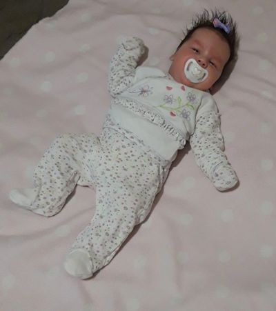 Baby in white floral pajamas lying on a pink polka-dot blanket with a pacifier.