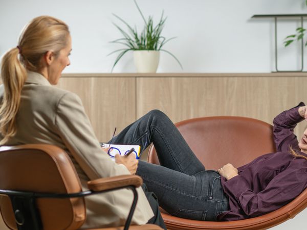 A woman lies on a couch talking to a therapist taking notes.