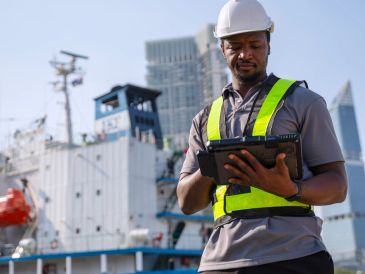Worker in safety gear using a tablet near a ship.