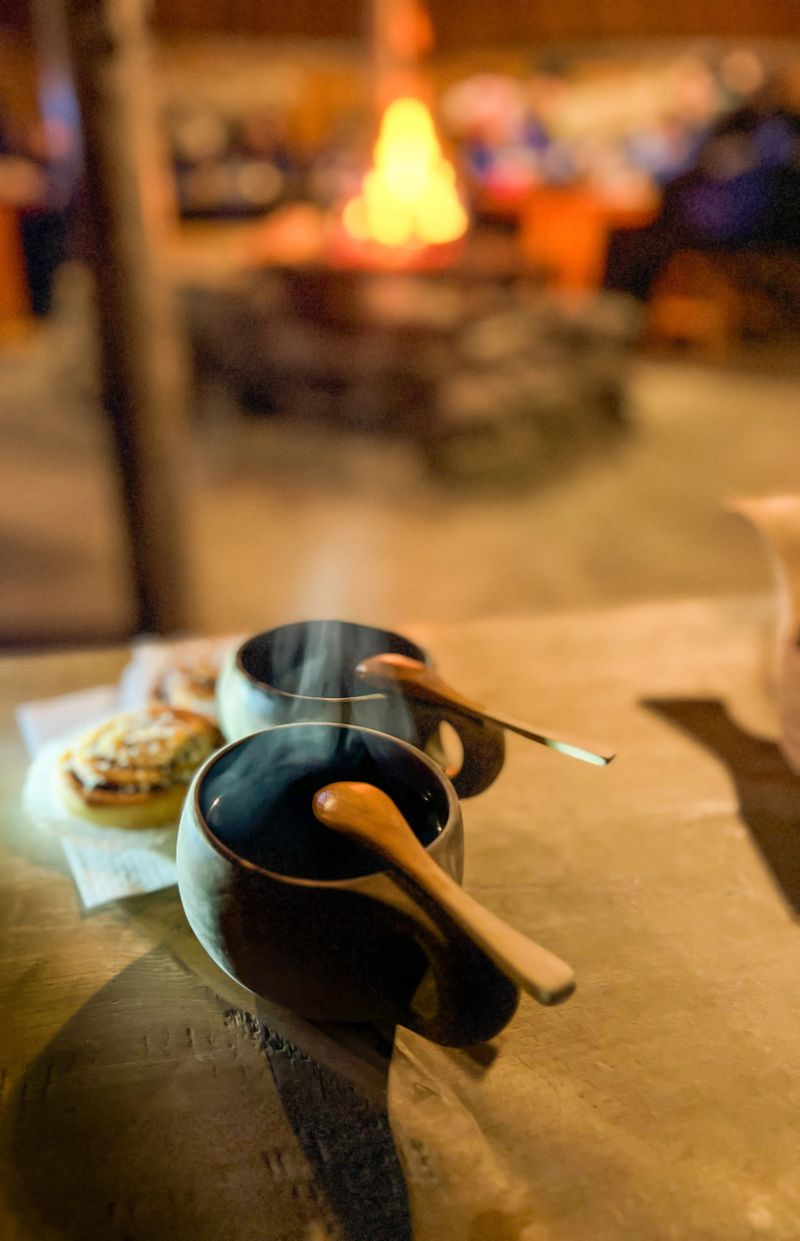 Two ceramic cups of hot red current drink and cinnamon bun with wooden spoons on a rustic wooden table, with a glowing fireplace in the background. Located in Lapland, Finland.