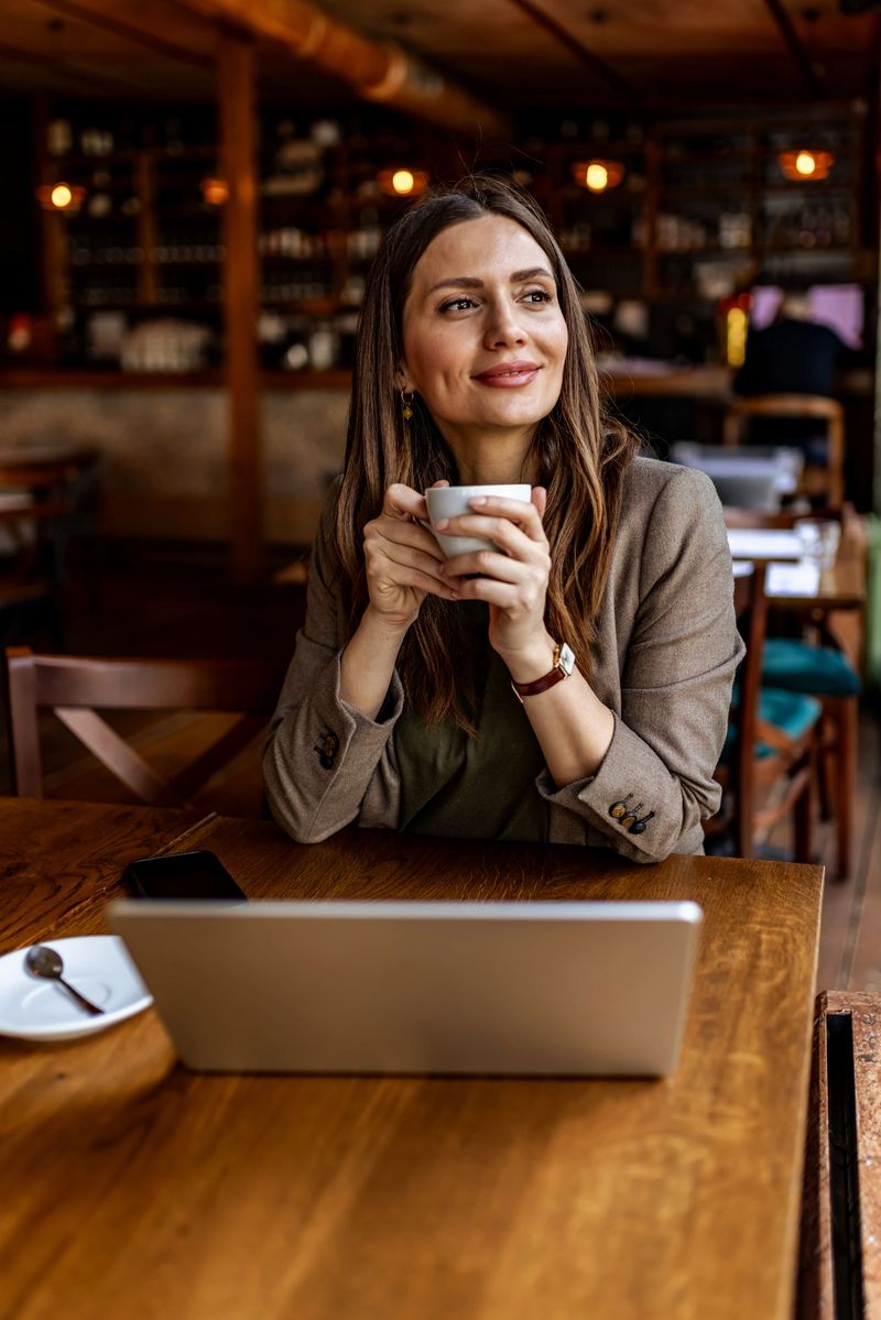 Professional woman sits in a cozy café holding a cup of coffee while working on a laptop, looking thoughtful and relaxed during a morning remote work break in a warm, modern workspace.