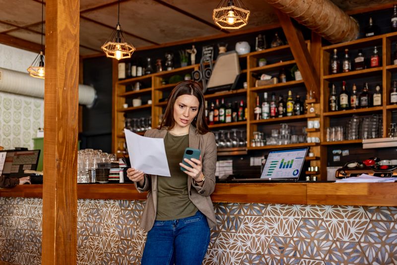 A small business owner in a rustic bar checks financial reports on her phone while holding invoices, with a laptop showing charts on the counter, conveying focus and concern about finances.