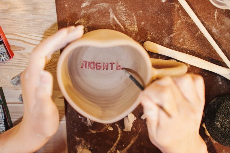Two hands work on a clay mug, adding the word Love inside it. Tools and materials are spread across a work surface in a pottery studio.