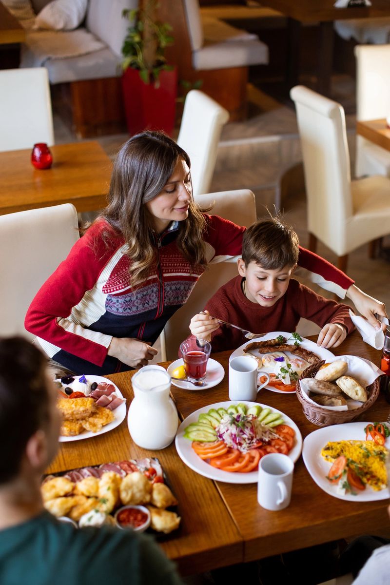 A family shares a meal at a restaurant. The table is filled with various dishes. A woman sits beside a boy who smiles while eating. Other family members are present, enjoying the food.