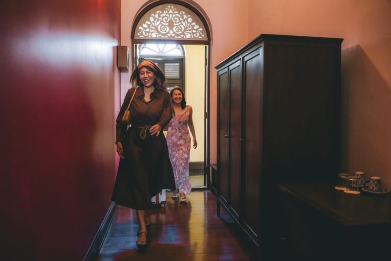 Two Asian adult women enter a heritage-style hotel room together while rolling a suitcase, capturing a welcoming travel arrival moment in a warm, elegant interior. Bangkok