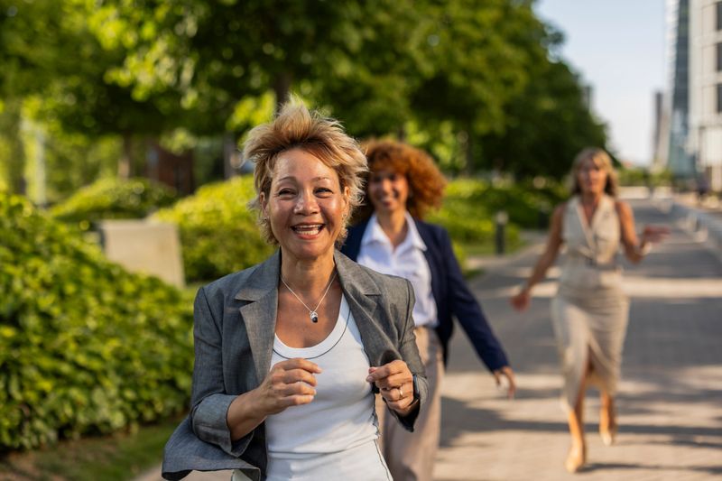 Group of diverse businesswomen running outdoors with big smiles, showcasing joy, success, and an active healthy lifestyle in a dynamic urban environment