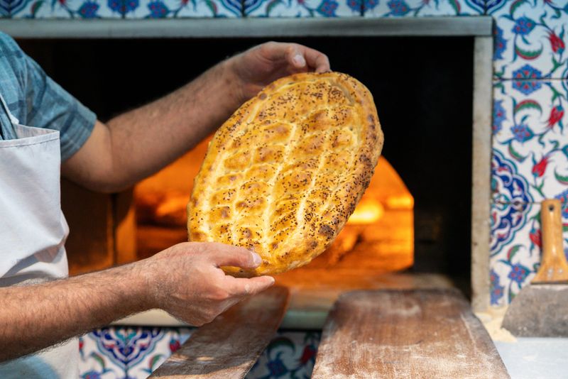 Close-up of a baker presenting freshly baked traditional Turkish Ramadan pide in front of a glowing wood-fired stone oven. The golden, hand-scored flatbread topped with sesame and nigella seeds represents centuries-old Ottoman baking tradition and is a cultural symbol of Ramadan and iftar meals in Turkey. Warm firelight, artisan craftsmanship, and authentic Middle Eastern culinary heritage create a rich, atmospheric food scene.