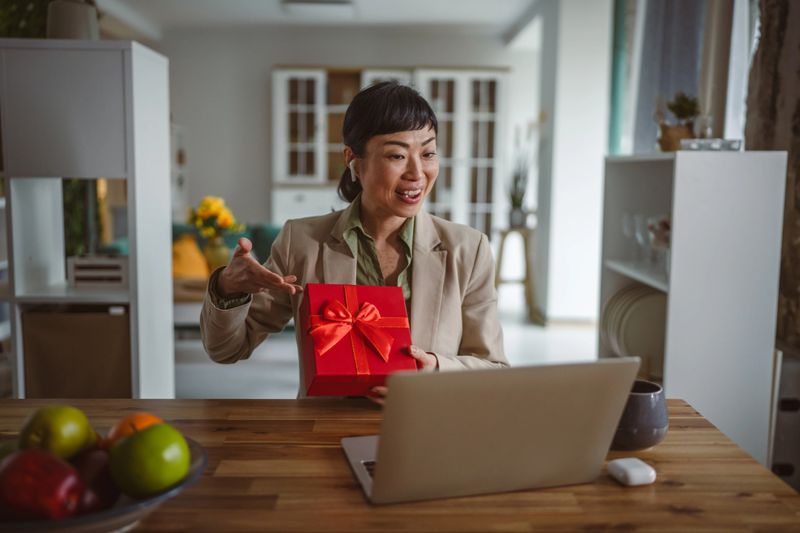 Woman feeling happy opening a red gift box while communicating with remote family or friends through a laptop video call, celebrating a special occasion