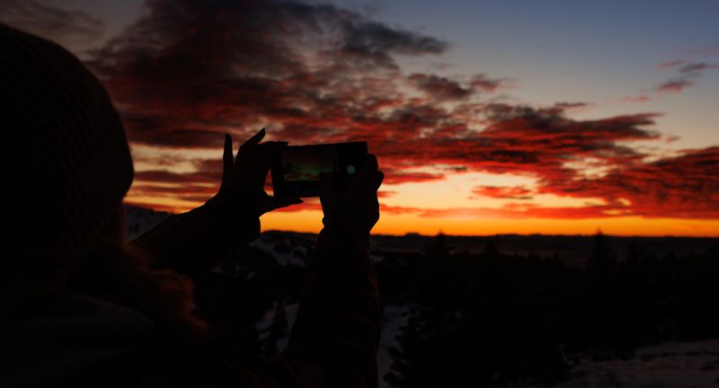 A person stands at a mountain overlook, holding a smartphone to capture the colorful sunset. The sky shows shades of orange and red while trees are visible in the foreground.