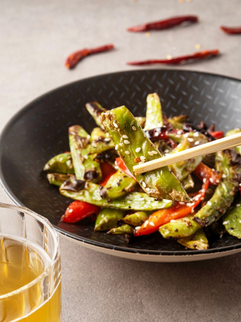 Close-up of chopsticks holding a spicy wok-fried green bean over a bowl with Kombucha.