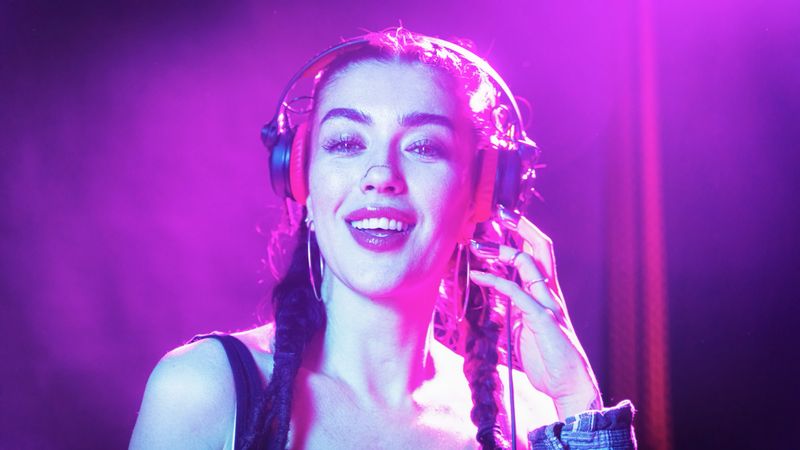 A woman stands in a club with headphones on, smiling as she DJs. Bright lights shine around her. The atmosphere feels lively and energetic.
