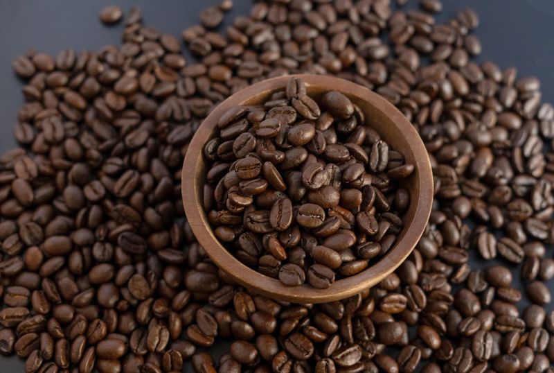 Top down view of aromatic roasted coffee beans filling a small brown wooden bowl, with many beans scattered around on a dark textured surface.